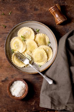 Seramik tabakta servis edilen ev yapımı ravioli makarnası rendelenmiş parmesan peyniri taze otlar kırsal ahşap arka planda peçeteyle çatallanıyor.