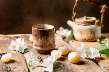 Seramik çaydanlık ve fincan, beyaz yasemin çiçekleri ve geleneksel Japon mochi şekerleriyle süslenmiş ahşap masa üzerinde huzurlu çay seremonisi atmosferi yaratıyor.