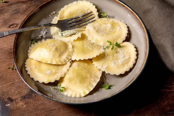 Seramik tabakta servis edilen nefis ev yapımı ravioli makarnası rendelenmiş parmesan peyniri taze otlar ve ahşap arka planda çatal.