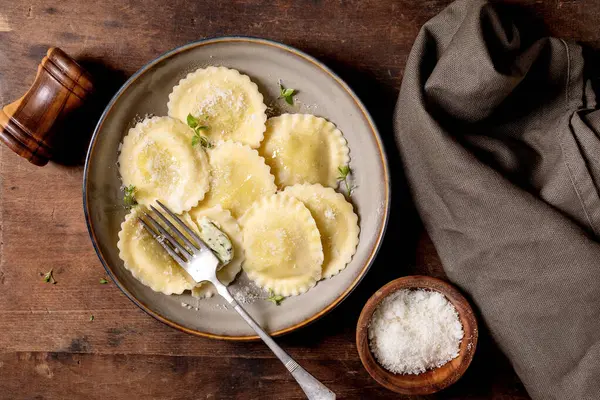 Seramik tabakta servis edilen ev yapımı ravioli makarnası rendelenmiş parmesan peyniri otları ve kırsal ahşap masa manzaralı çatal.