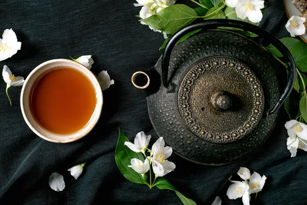 Geleneksel dökme demir çaydanlık, koyu yeşil keten arka planda beyaz yasemin çiçekleriyle çevrili. Rahatlama refahı için üst görünüm düz yatay bileşimi