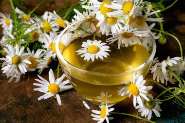 Cam bardak papatya çayı ahşap arka planda taze papatya çiçekleriyle çevrili. Doğal çiçekli bitkisel içecek huzur dolu bir sağlık ortamı yaratır..