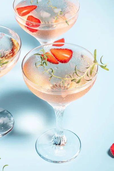 Glasses with cold pink champagne or punch with wine strawberries, ice and rosemary on blue background.