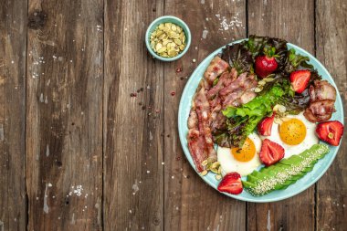 Plate with fried eggs, bacon, avocado, strawberries and fresh salad on a dark background. Ketogenic diet food, healthy meal concept, Long banner format. top view.