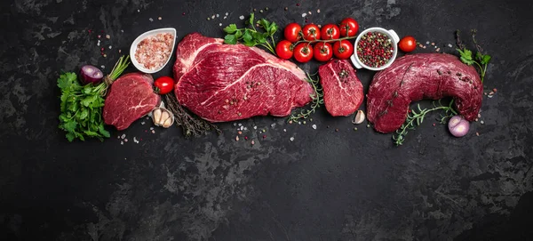 Assortment of raw cuts of raw beef meat steaks with spices on a dark background. Long banner format. top view.