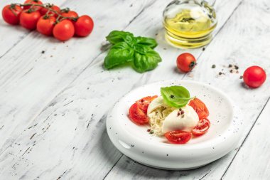 Fesleğenli kremalı İtalyan Burrata peyniri, zeytinyağı ve beyaz tabakta beyaz ahşap zemin üzerinde kiraz domatesleri. afiş, menü, metin için tarif yeri, üst görünüm.