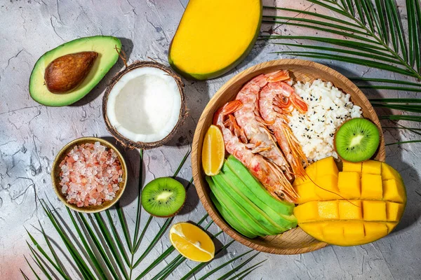 tropical bowl with avocado, prawns, rice, mango, kiwi and coconut, tropical food on the sea beach on light background with tropical leaves. Healthy food, clean eating, Buddha bowl, top view,