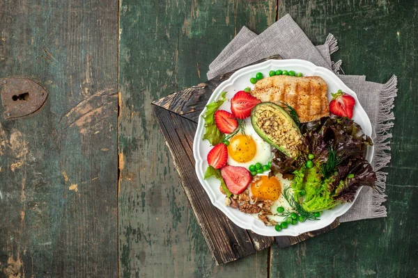 Plate with keto diet food. Keto breakfast Fried egg, avocado, strawberry, grilled chicken fillet, nuts and fresh salad, Ketogenic diet. Healthy food concept, top view.