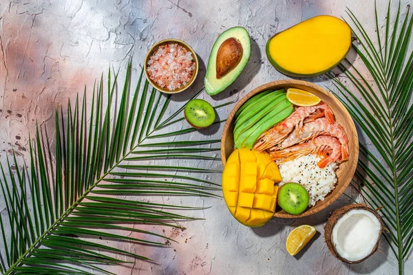 tropical bowl with avocado, prawns, rice, mango, kiwi and coconut, tropical food on the sea beach on light background with tropical leaves. Healthy food, clean eating, Buddha bowl, top view,