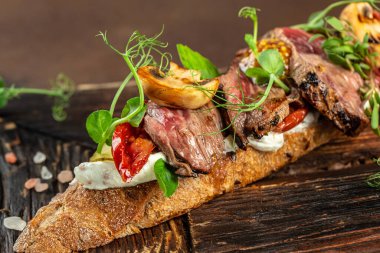 Biftekli sandviç, dilimlenmiş biftek, mantar, kurutulmuş domates, salatalık turşusu ve koyu arkaplanda yeşillikler. Üst Manzara. metin için yer,