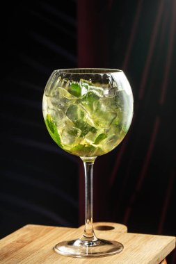 Nane yaprakları ve buz küpleriyle yeşil karışımı, ahşap bir tahtanın üzerinde, kapkara bir zemine karşı büyük bir bardakta servis edilir..