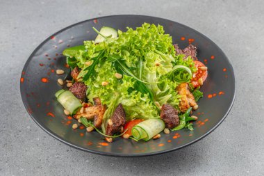 Izgara etli taze yeşil salata, salatalık, vişneli domates ve kırmızı soslu siyah tabakta servis edilen kavrulmuş çam fıstığı..