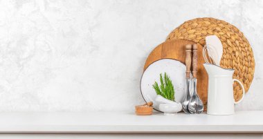 Ahşap ve seramik kaplar, dokunmuş plasemat, beyaz sürahi, tuz, şifalı otlar ve beyaz bir masa üzerinde düzgün bir şekilde dizilmiş aletlerle minimalist bir sade mutfak düzeneği..