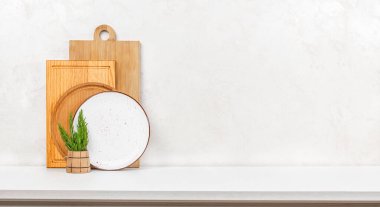 Kesme tahtaları, seramik plaka ve beyaz tezgahın üzerinde dokulu bir ışık duvarına karşı kırsal bitki tutacağı olan temiz bir minimalist mutfak görüntüsü..