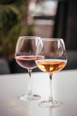 Beyaz bir yüzeyde kırmızı ve kehribar şarapla dolu iki zarif şarap kadehi, rahat bir ortamda yumuşak bir iç mekan ışığını yansıtıyor..