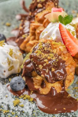 Çikolata yağmuruna tutulmuş zengin bir profiterol krema, taze çilek, yaban mersini, nane yaprağı ve ezilmiş fındıkla süslenmiş..