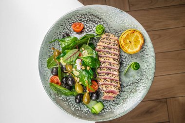 Taze ıspanak ve zeytin salatası, domates, çam fıstığı, salatalık sarmalı ve ızgara limon ile servis edilen susam kabuklu kurutulmuş ton balığı dilimli güzel bir tabak..