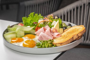 Renkli ve dengeli bir kahvaltı tabağı sunar. Yumurta, jambon, peynir, taze sebze, zeytin, edamame ve yuvarlak bir tabakta güzelce hazırlanmış kızarmış ekmek..