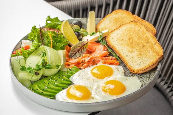 Canlı ve besleyici bir kahvaltı tabağı, güneşli yumurta, tütsülenmiş somon, avokado, salatalık kurdelesi, peynir, taze yeşillik, zeytin, limon ve kızarmış ekmek..