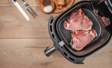 Çiğ domuz pirzolası, mutfak aletleri ve baharatlarla çevrili, ahşap bir tezgahta, uygun ve sağlıklı bir pişirme işlemi için hazır bir şekilde, bir hava kızartma sepetinin içine yerleştirilir..