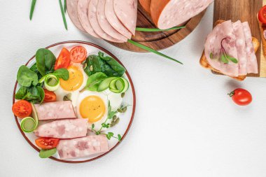 Sağlıklı bir kahvaltı tabağı; güneşli yumurta, meze sosisi dilimleri, kiraz domatesleri, salatalık kurdeleleri ve taze yeşillikler canlı bir sabah yemeği için beyaz arka planda servis edilir..