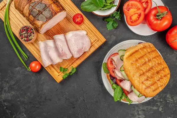 Taze dilimlenmiş jambon sebze ve otlarla birlikte bir kesme tahtasında servis edilirken, jambon, domates, salatalık ve yeşillik dolu bir sandviç de yakınlardaki bir tabakta duruyor..