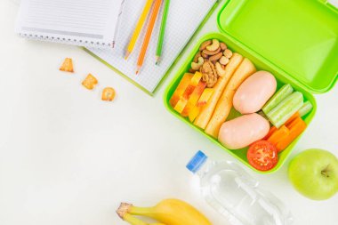 Sebzeler, peynirler, sosisler, fındıklar, meyveler ve defterlerin ve renkli kalemlerin yanına yerleştirilmiş bir şişe suyla dolu yeşil bir beslenme çantasında sağlıklı bir okul yemeği..