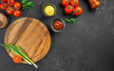 Koyu arkaplanda yeşil soğan ve domatesli yuvarlak ahşap kesme tahtası. Taze malzemeler ve baharatlarla çevrili, yemek hazırlığı için hazır..
