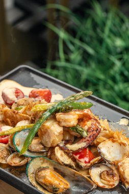 Karides, midye, midye, istiridye ve sebzeli cızırtılı deniz ürünleri karışımı kremalı ve otlu siyah bir ızgara tabağında sıcak servis edilir..