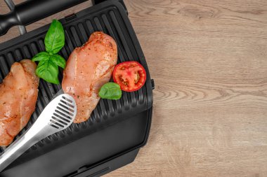 İki çiğ, terbiye edilmiş fesleğen yapraklı tavuk göğsü ve bir domates dilimi ahşap bir yüzeydeki metal maşanın yanına elektrikli ızgaraya yerleştiriliyor..