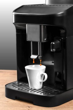 Siyah, modern bir kahve makinesi, beyaz bir fincana espresso demler. Ahşap bir tezgahın üzerine, parlak bir arka planı olan bir fincana koyar..
