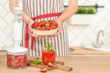 Çizgili önlüklü bir insan, ahşap mutfak tezgahında blender dolusu çilek ve bir bardak çilekli smoothie bulundurur...