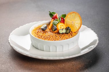 Karamelleştirilmiş şeker hamurlu klasik creme brulee taze böğürtlenler, nane yaprakları ve dekoratif portakal dilimleriyle süslenmiştir..