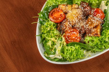 Taze yeşil salata, ızgara patlıcan, kiraz domatesleri ve tahtadan bir masada servis edilen beyaz oval kasede susam tohumları..