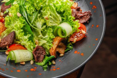 Koyu bir tabakta servis edilen taze yeşil salata, ızgara sığır eti, salatalık, kiraz domatesleri, mantarlar ve çam fıstığı..