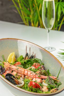 Karides, midye ve taze yeşilliklerle dolu canlı bir deniz ürünleri salatası, serinletici bir bardak köpüklü şarapla birlikte kırsal bir kasede servis edilir..