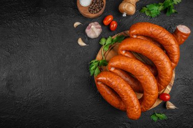 Ahşap bir tahtanın üzerinde tütsülenmiş sosis, otlar, sarımsak, biber ve domatesler koyu dokulu arka planda. Üst Manzara. Boşluğu kopyala.