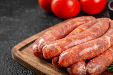 Arka planda olgun kırmızı domateslerle ahşap kesim tahtasına istiflenmiş pişmemiş domuz sosislerinin yakın plan görüntüsü..