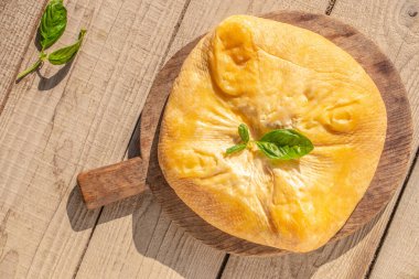 Tütsülenmiş peynirin tamamı fesleğen yaprağıyla süslenmiş doğal bir kabuğa sarılmış. Sıcak, doğal bir ışıkla kırsal bir ahşap tahtada servis ediliyor. Üst Manzara. Boşluğu kopyala.