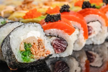 Renkli suşi rulolarının çeşitli çeşitleri parlak siyah bir yüzey üzerinde sergilenmekte, canlı malzemeler ve taze malzemeler sergilenmektedir..