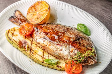 Beyaz bir tabakta ızgara balık, kavrulmuş kabak, kiraz domatesleri ve limonla servis edilir. en üstteki görünüm.