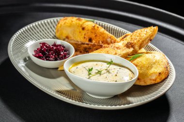 Kremalı ezme ve tatlı karamelli soğan reçeli ile servis edilen gevrek Focaccia ekmeği, gri oval tabakta taze otlarla sunuluyor..