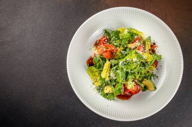 Taze yeşil salata roka, marul, avokado, kiraz domatesleri ve rendelenmiş peynir ile birlikte koyu bir arka plan üzerinde beyaz bir tabakta servis edilir..