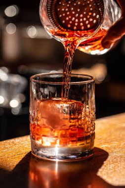 Sıcak ışık ve ahşap tezgahtaki yansımalarla vurgulanmış büyük bir buz küpüyle süzgeçten geçip bir bardağa dökülen klasik kokteyl..