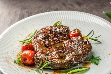 Kaba tuz, taze otlar ve beyaz tabakta kızartılmış domatesle süslenmiş ızgara biftek..