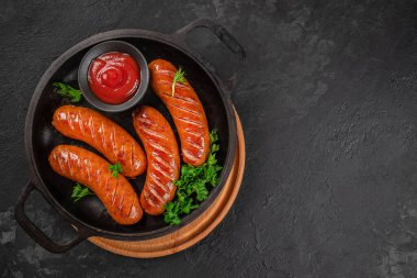 Maydanozlu ızgara sosis ve koyu arkaplanda servis edilen dökme demir tavada ketçap. Üst Manzara. Boşluğu kopyala.