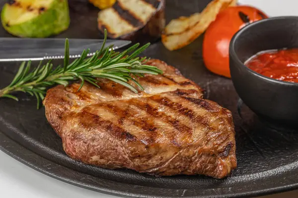 Biberiyeli ızgara biftek, ızgara sebze ve siyah tabakta servis edilen domates sosu..