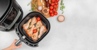 Raw chicken drumsticks with tomatoes and herbs in air fryer basket, preparation for healthy cooking at home, modern kitchen appliance top view with copy space.