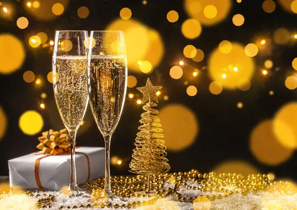 İki bardak köpüklü şampanya, şenlikli altın ışıklar, Noel ağacı süsü ve hediye kutusu. Kopyalama alanı olan tatil kutlaması arkaplanı.