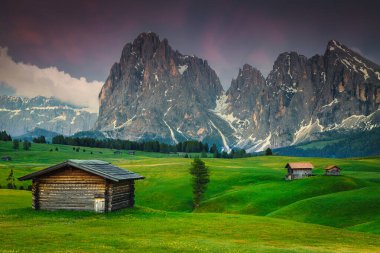 Yeşil tarlada küçük şirin ahşap kulübeleri ve günbatımında karlı dağları olan muhteşem yaz manzarası, Alpe di Siusi, Dolomitler, İtalya, Avrupa